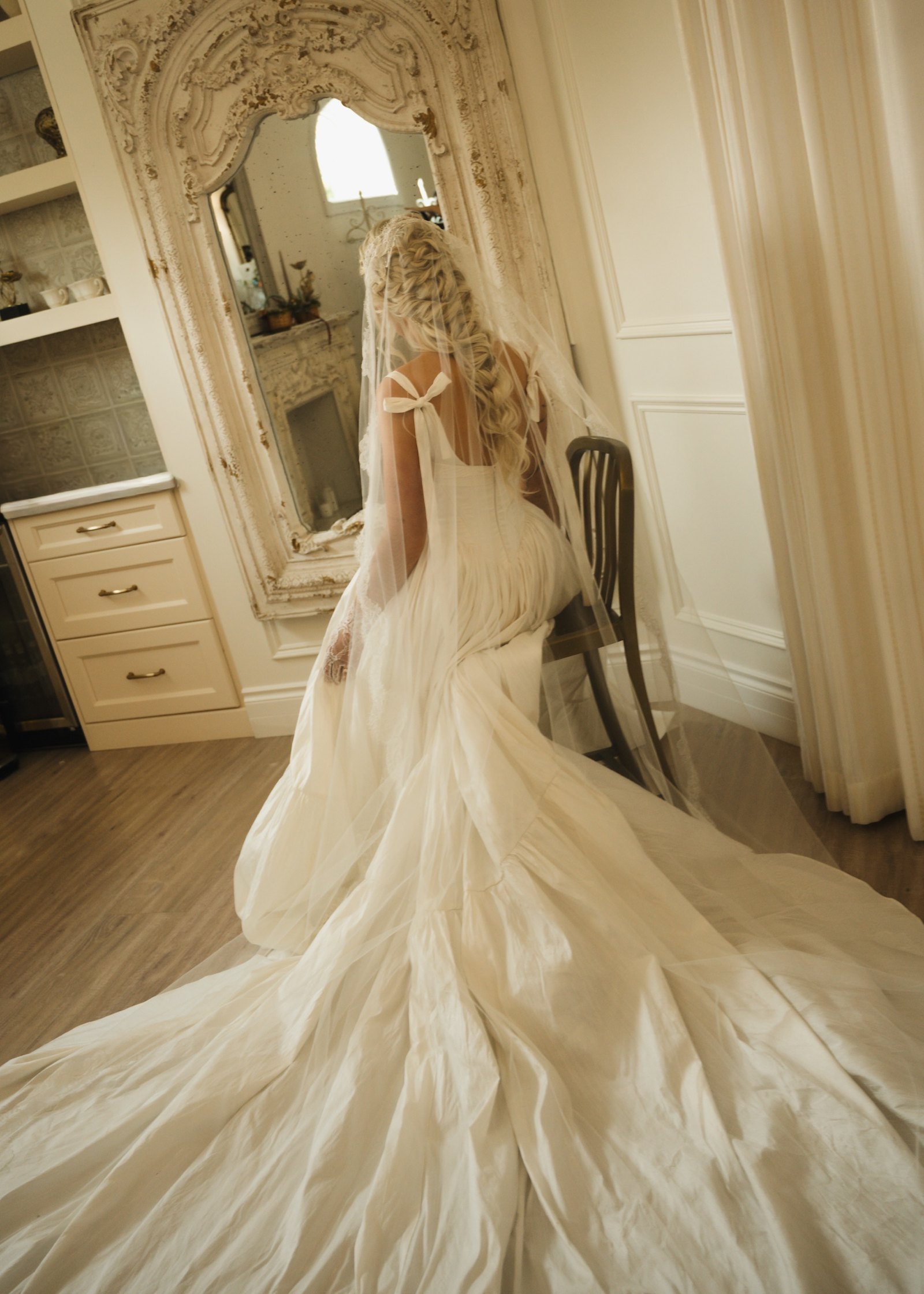 Bride in a lace-edged veil worn over a long textured braid, seated at a mirror while getting ready